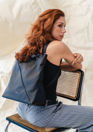 Woman sitting on a chair with a black backpack, wearing a black top and blue pants.